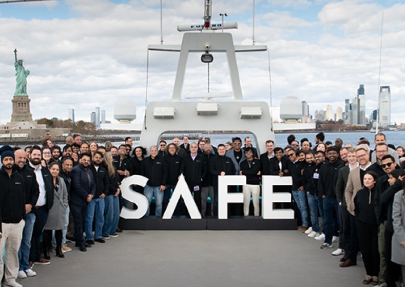 The SAFE team gathered around a SAFE sign on a boat in New York harbour, with the Statue of Liberty and the Manhattan skyline behind them.